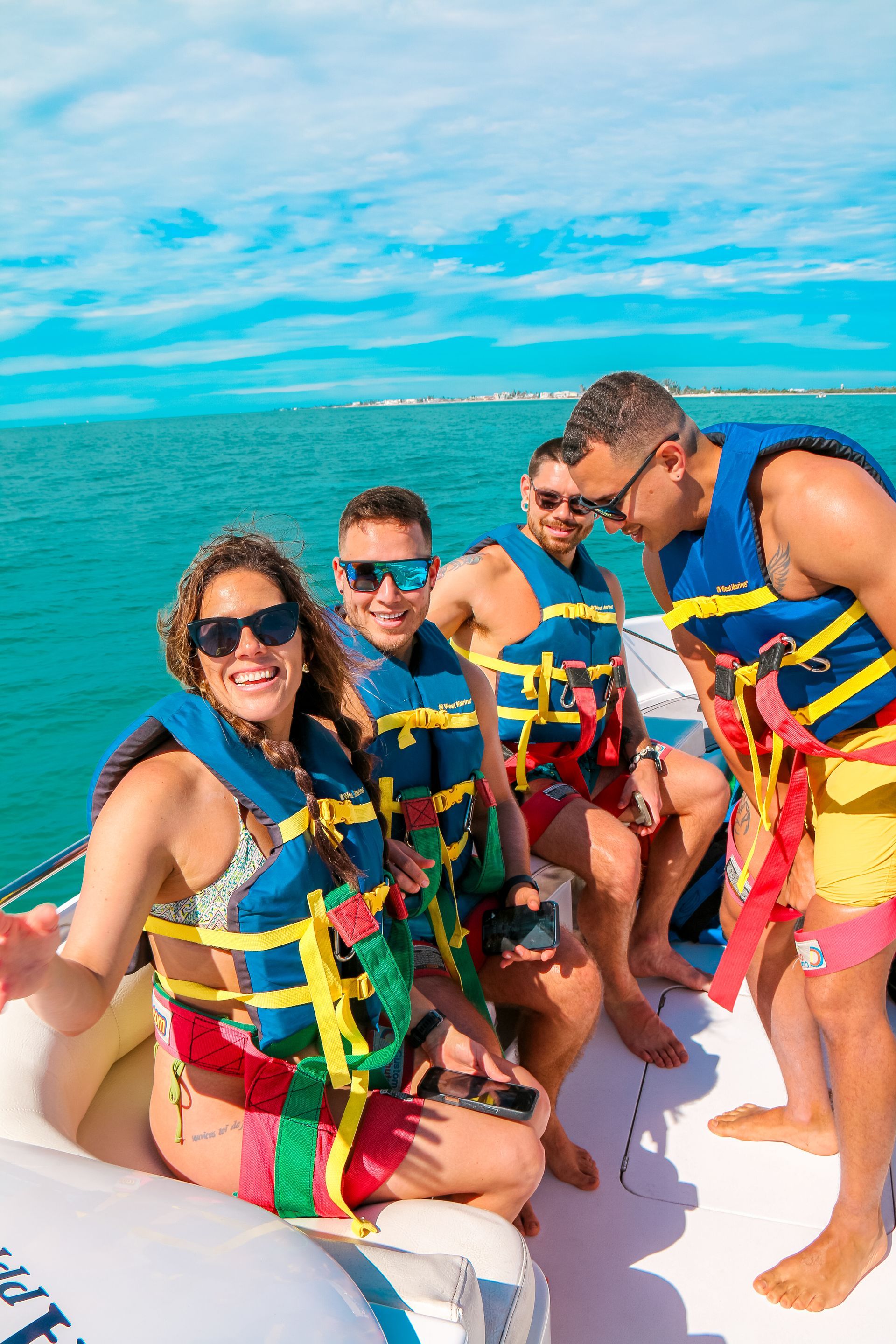 Four people on a boat, wearing life vests. Blue water and sky. Woman smiles, gesturing.