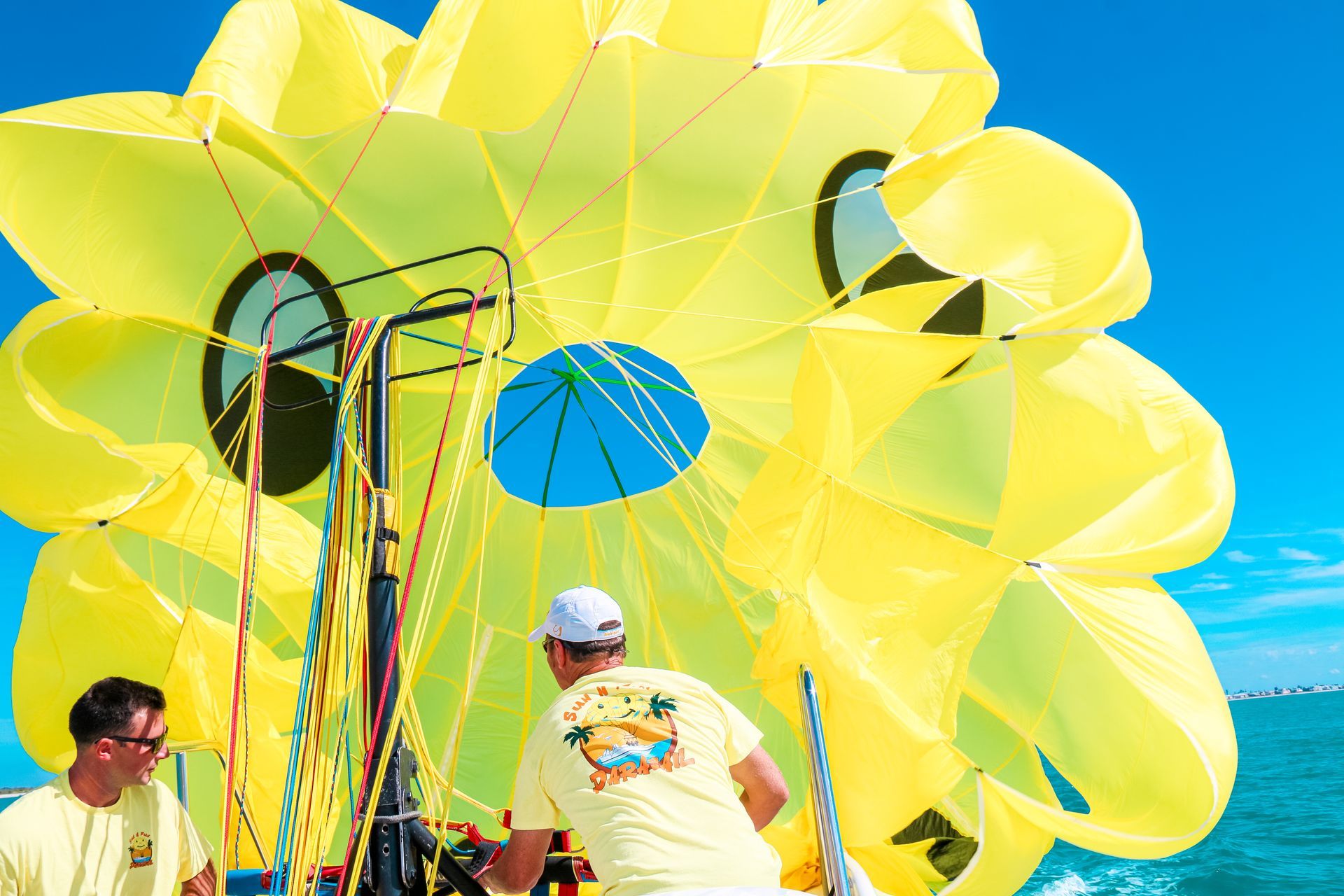 Yellow parasail being prepared by two men on a boat, blue sky backdrop.