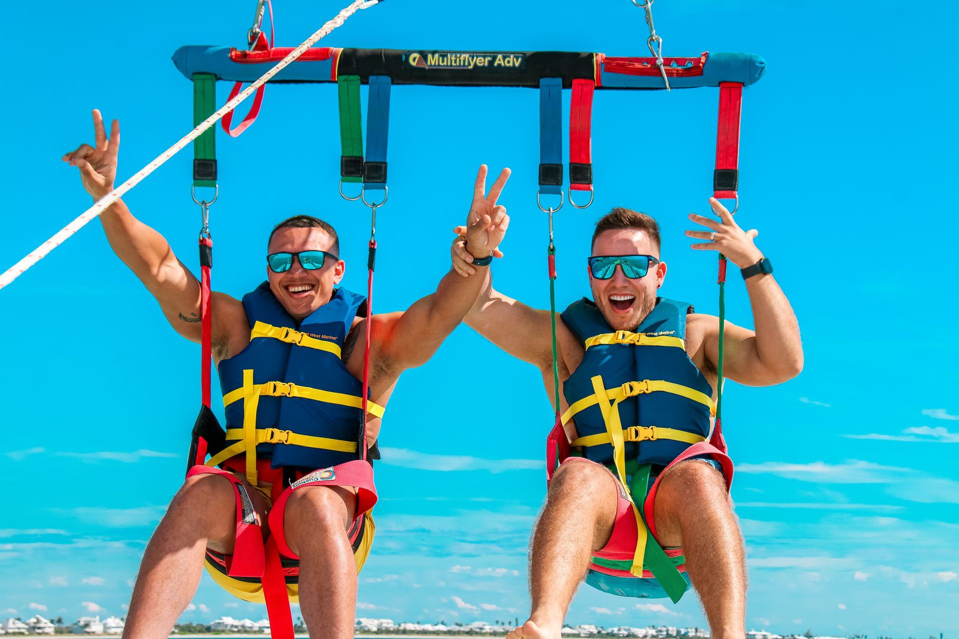 Two men parasailing, holding up peace signs, wearing life vests and sunglasses against blue sky.