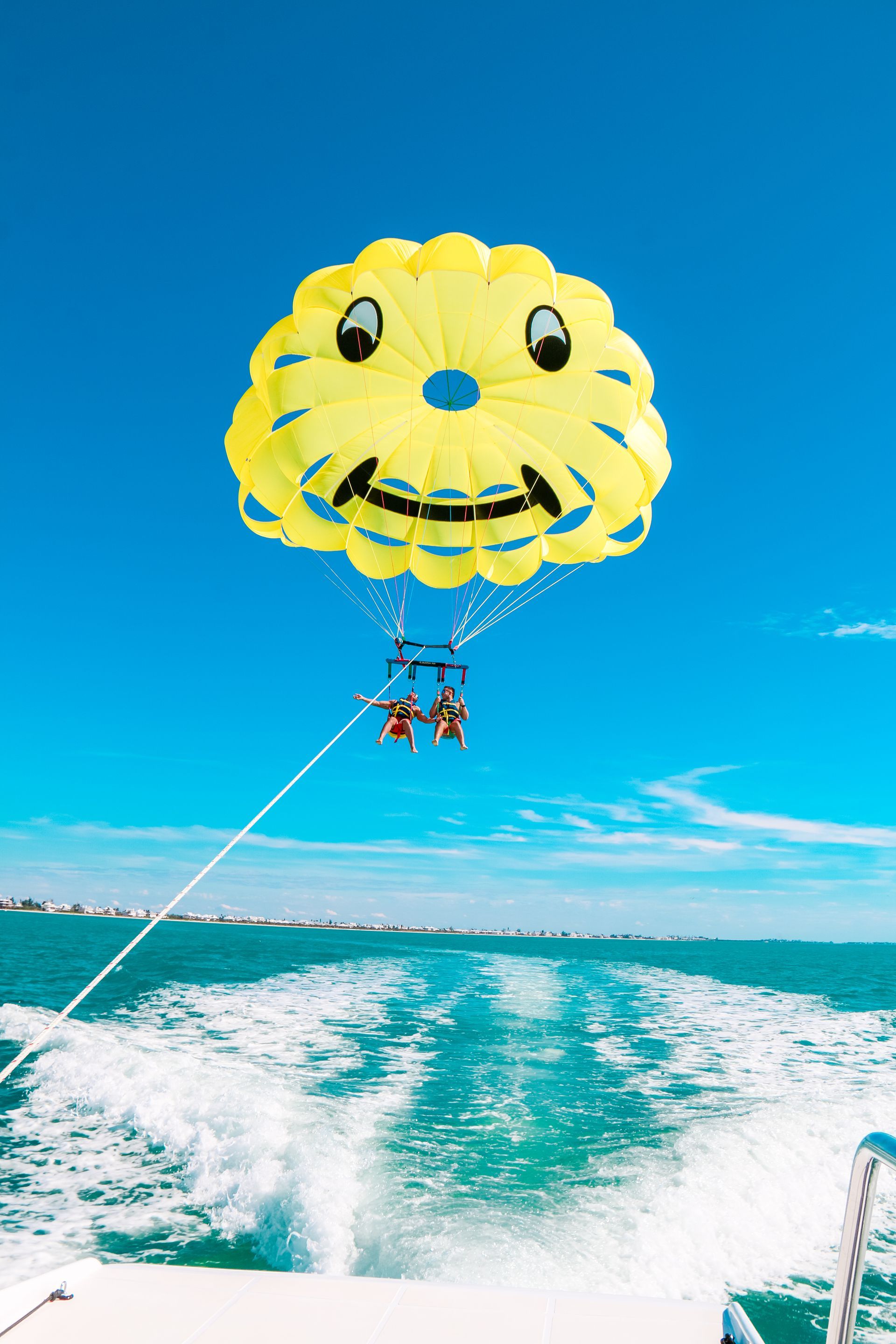 Yellow parasail shaped like a smiley face over ocean; two figures parasailing behind a boat.