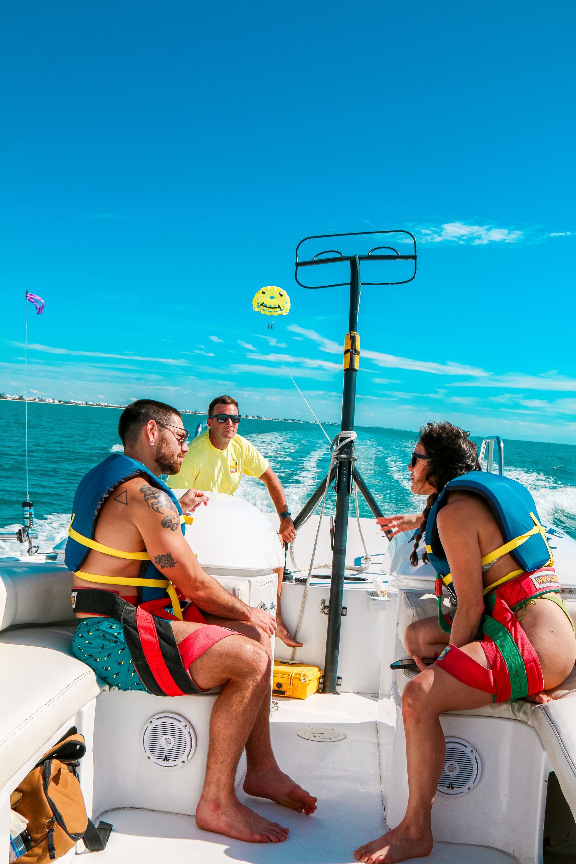 Couple on a boat wearing life vests, preparing to parasail. Blue sky and water, a crew member is present.