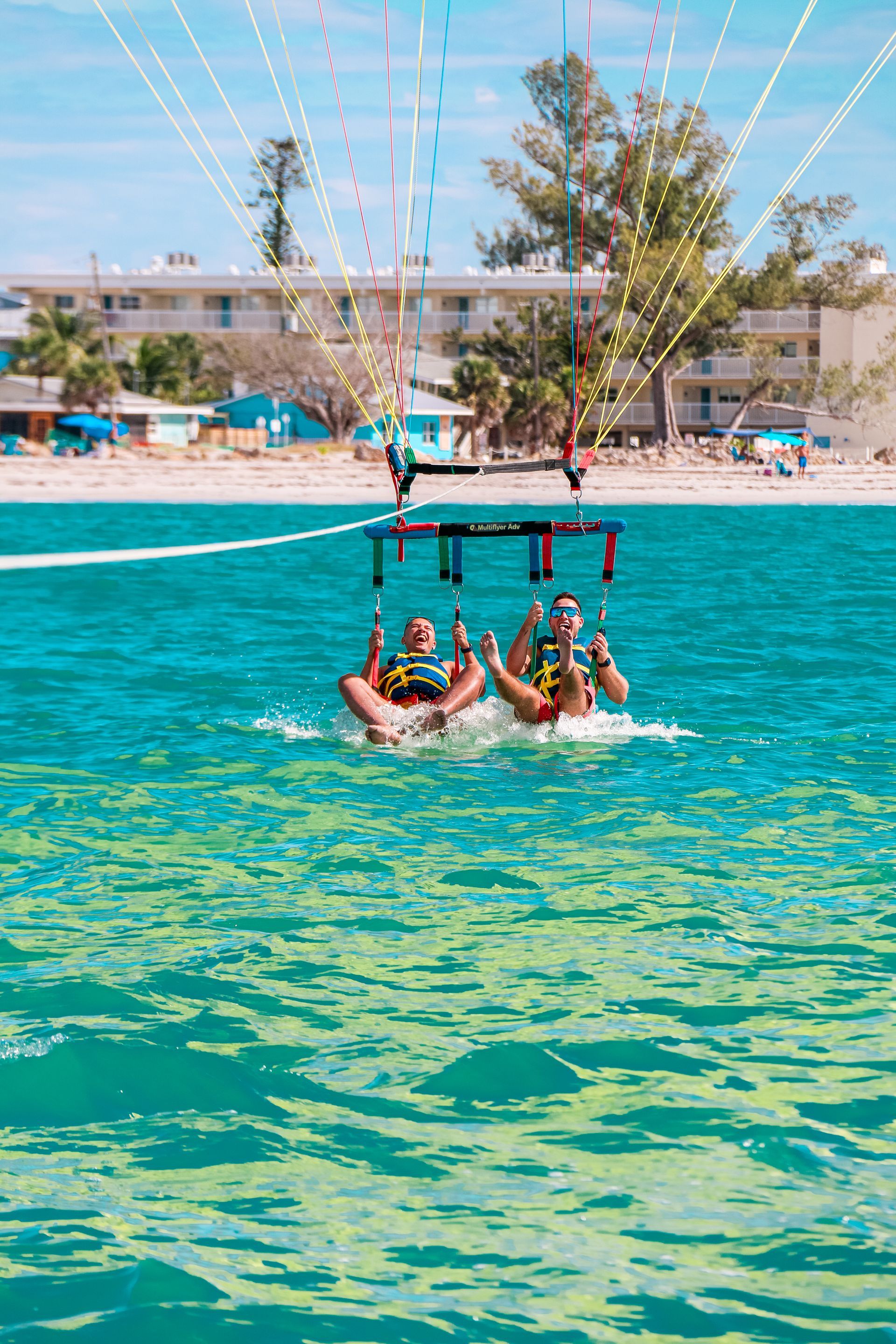 Two people parasailing above turquoise water near a beach with buildings.