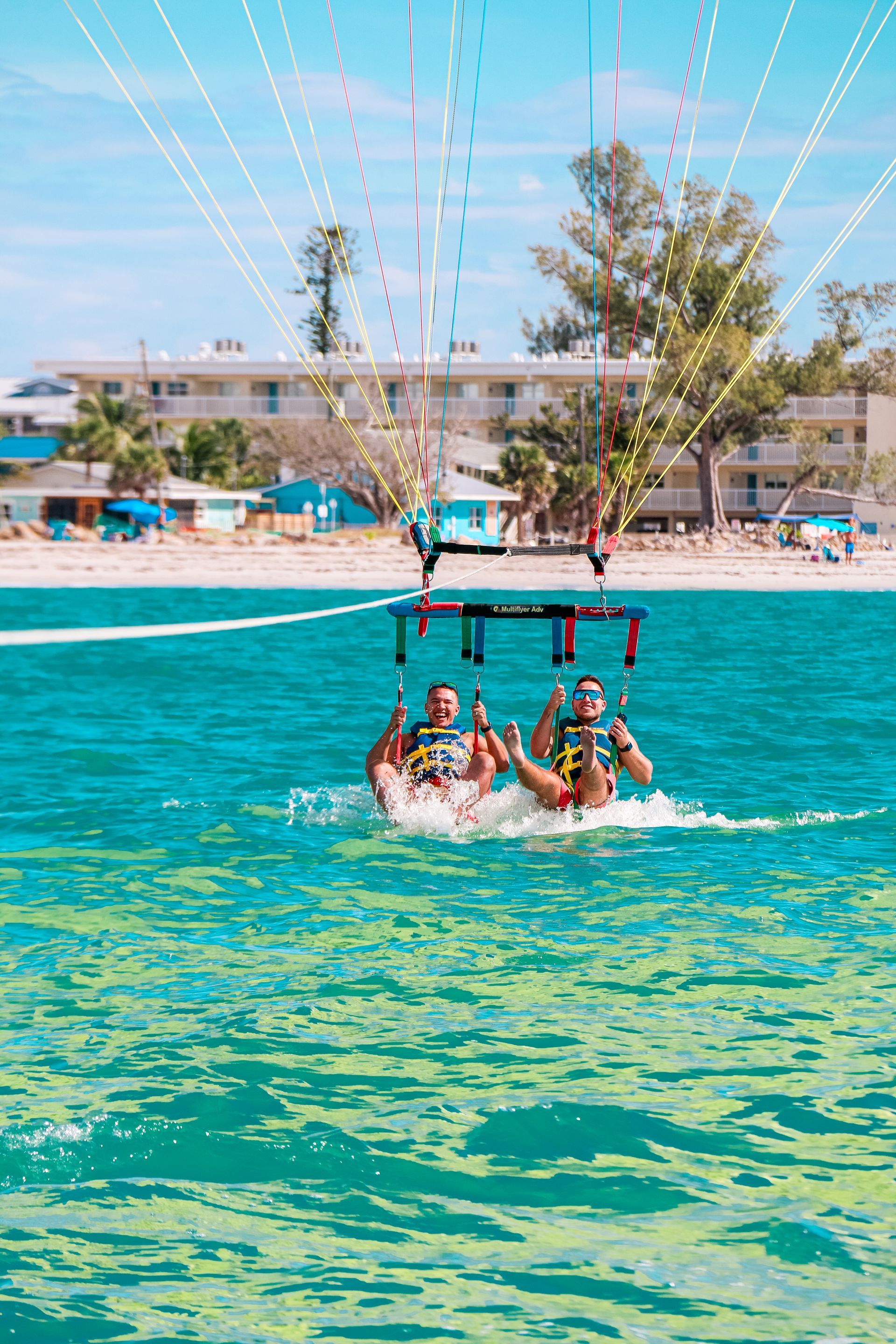 Two people parasailing over turquoise water near a beach with buildings.