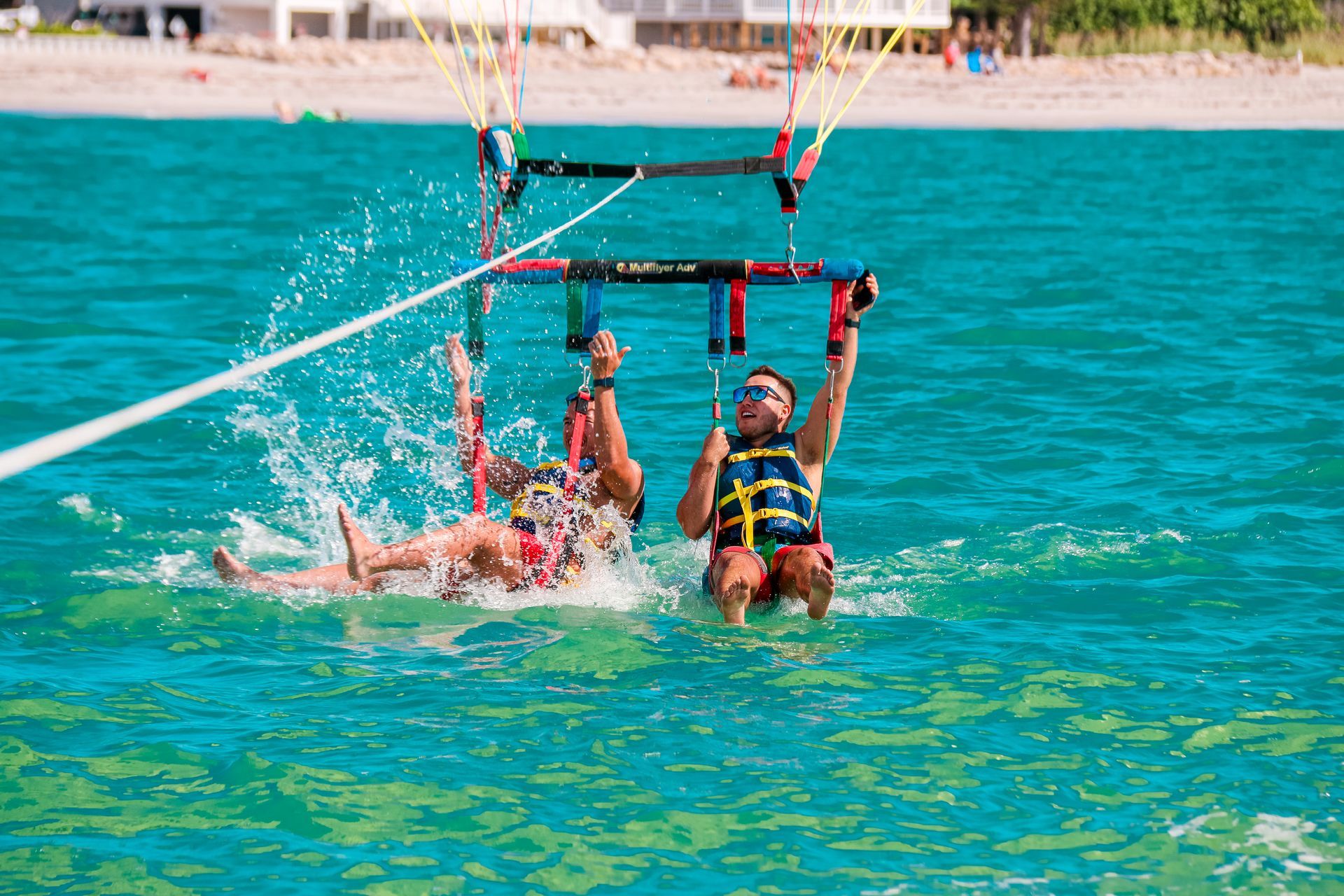 Two people parasailing, splashing into turquoise water near a beach.