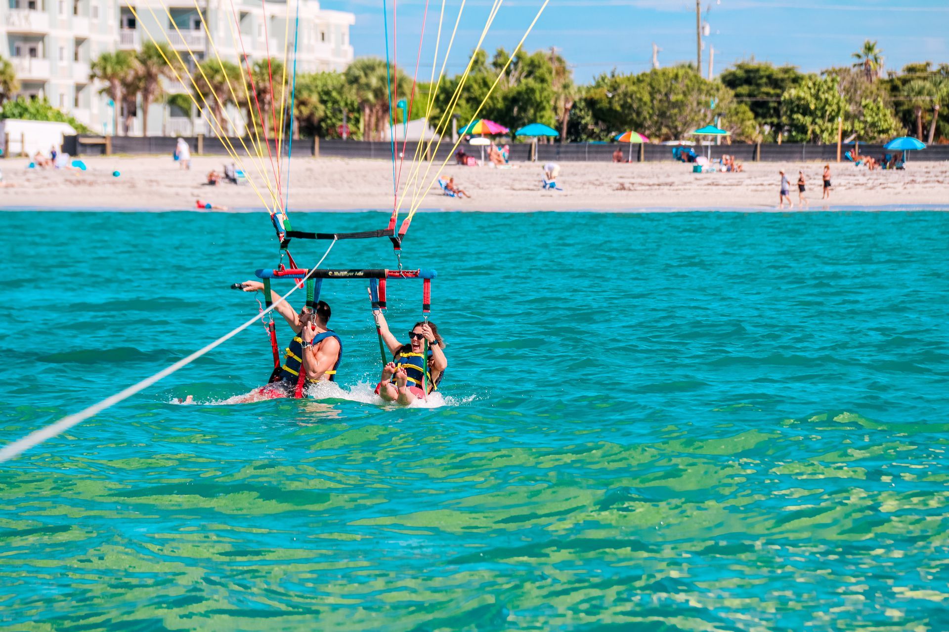 Two people parasailing over turquoise water, beach in the background.