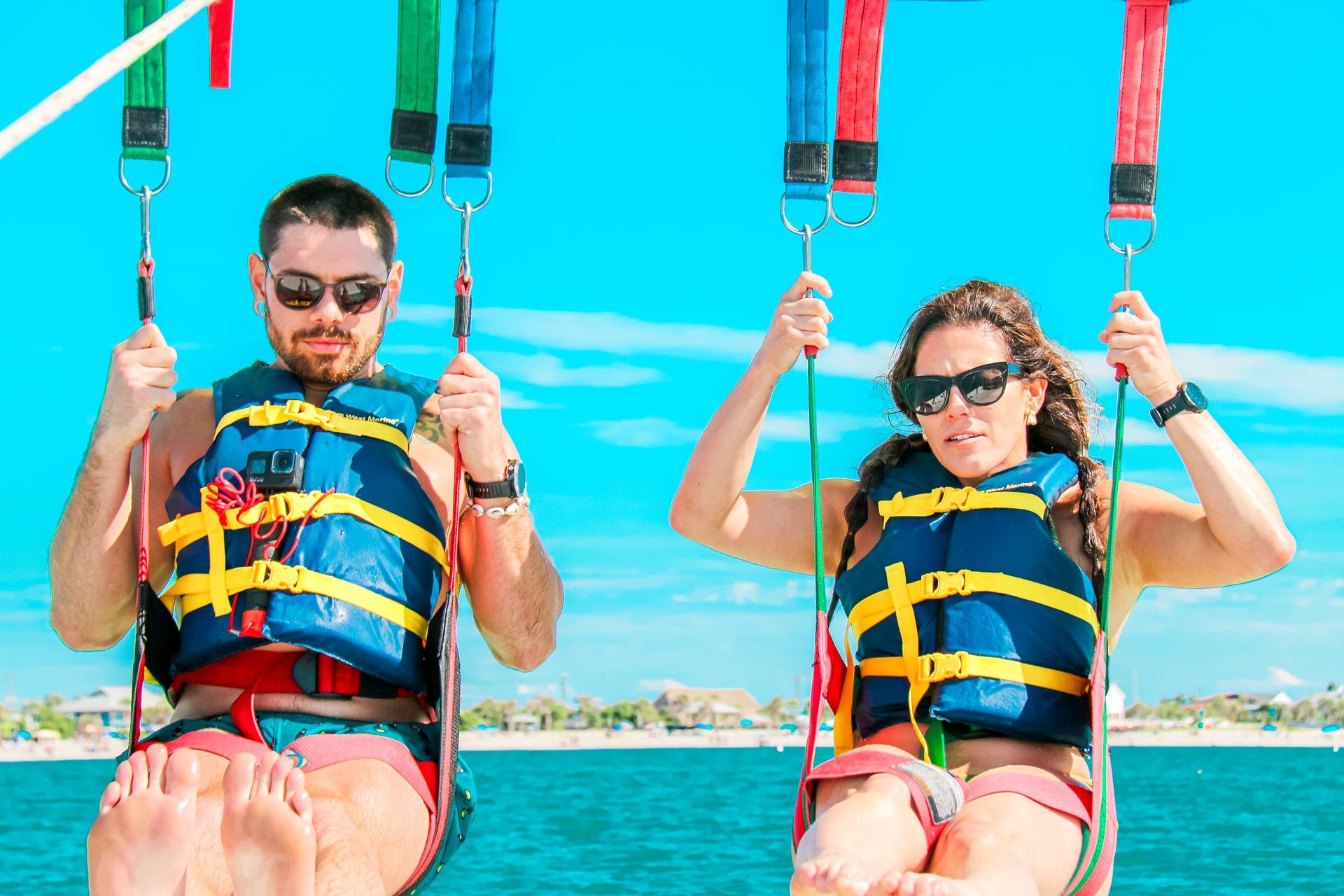 Two people parasailing above a beach with blue water and sky. They wear life vests and sunglasses.