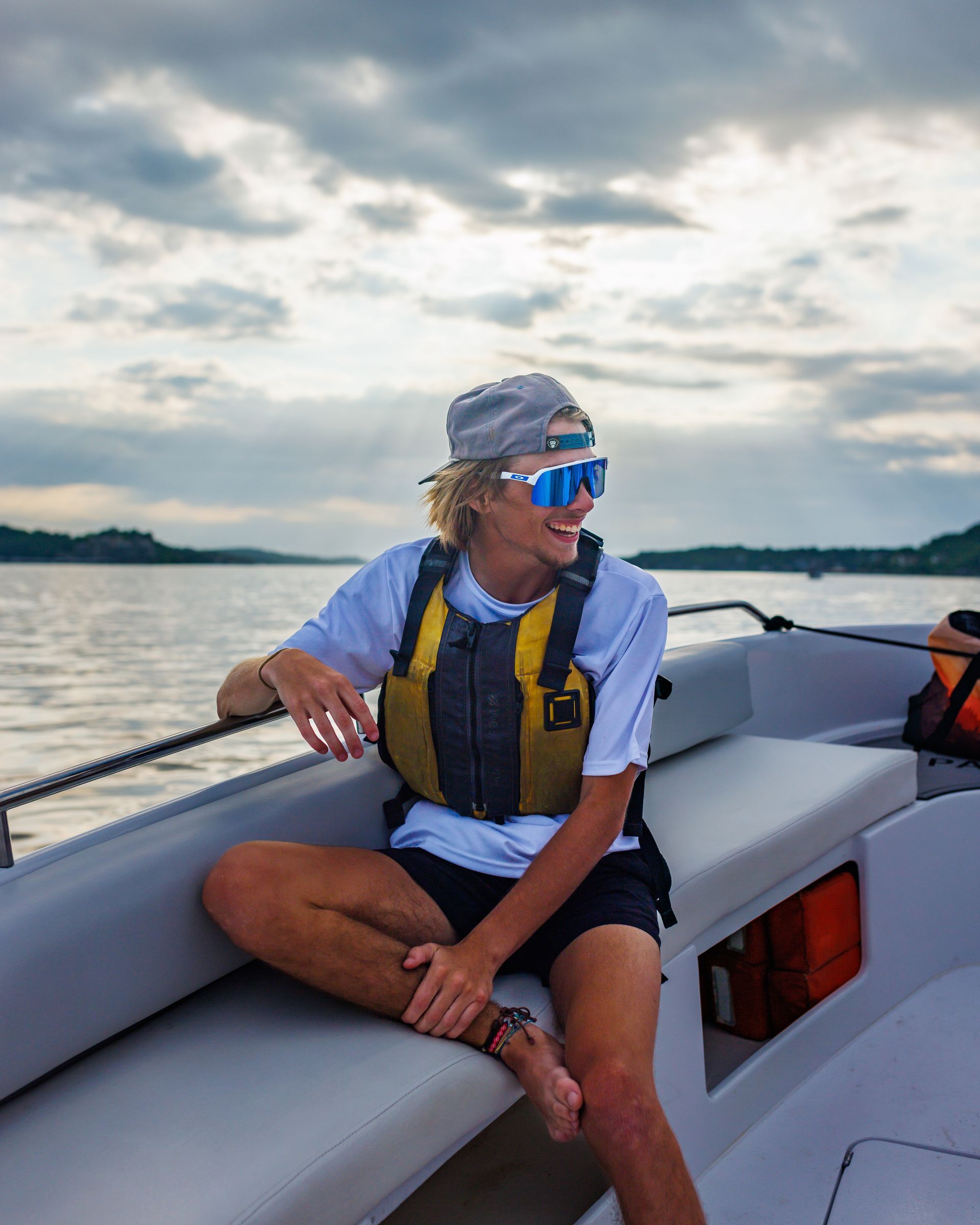 Person sitting on a boat, wearing sunglasses, a life vest, and a hat, smiling at the water.