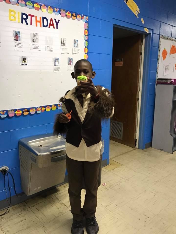 A young boy is standing in front of a birthday sign in a classroom.
