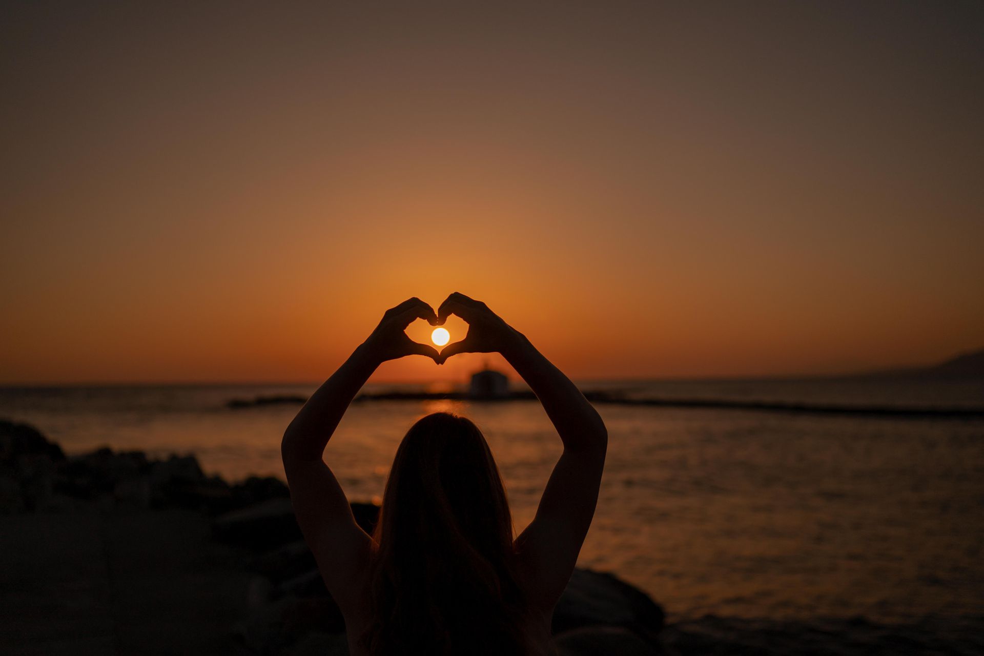 Eine Frau formt bei Sonnenaufgang vor der Sonne mit ihren Händen ein Herz.