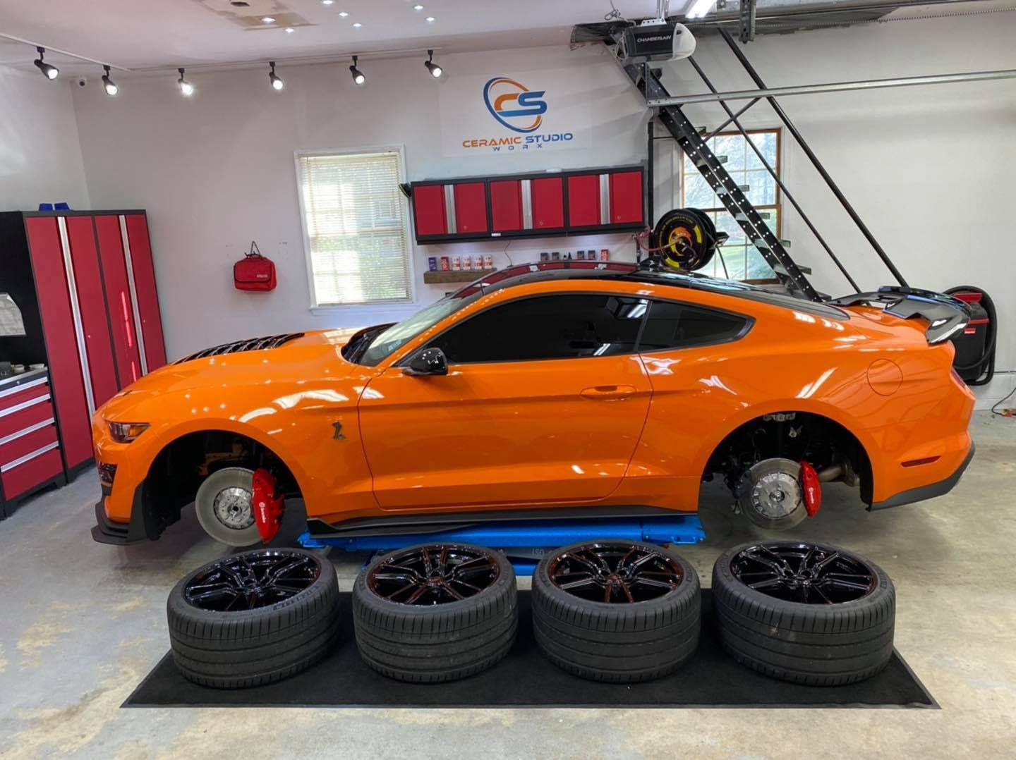 Orange Mustang Shelby in a garage, tires removed, four new black tires in front.