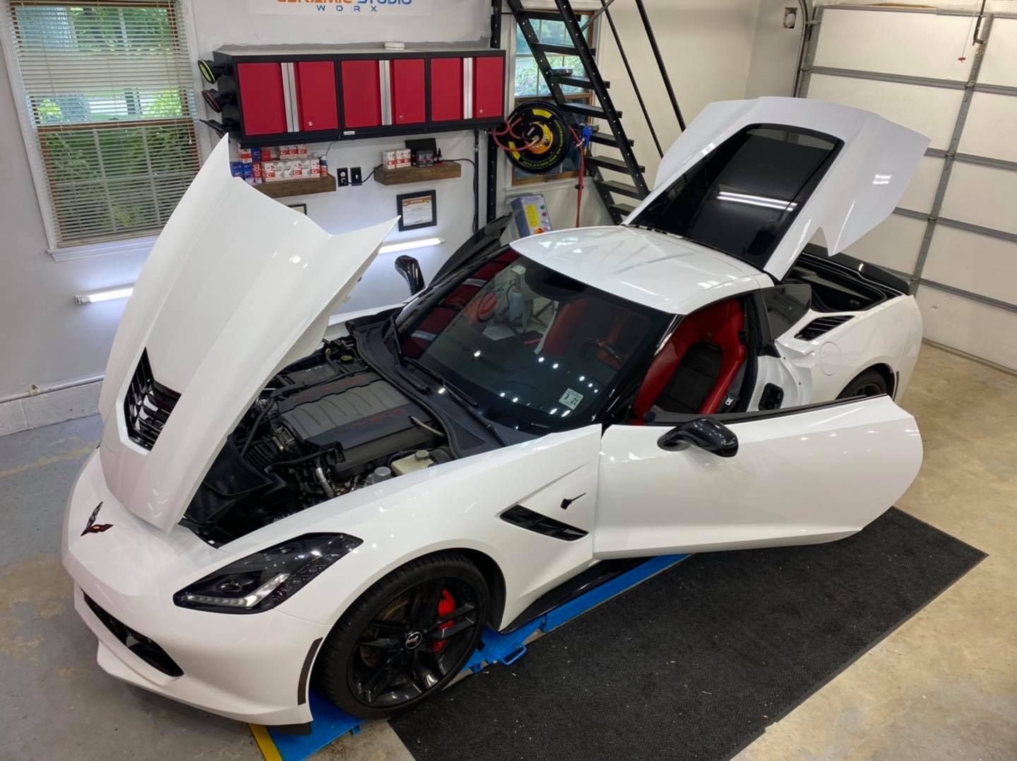 White Corvette convertible with open hood and door in a garage.