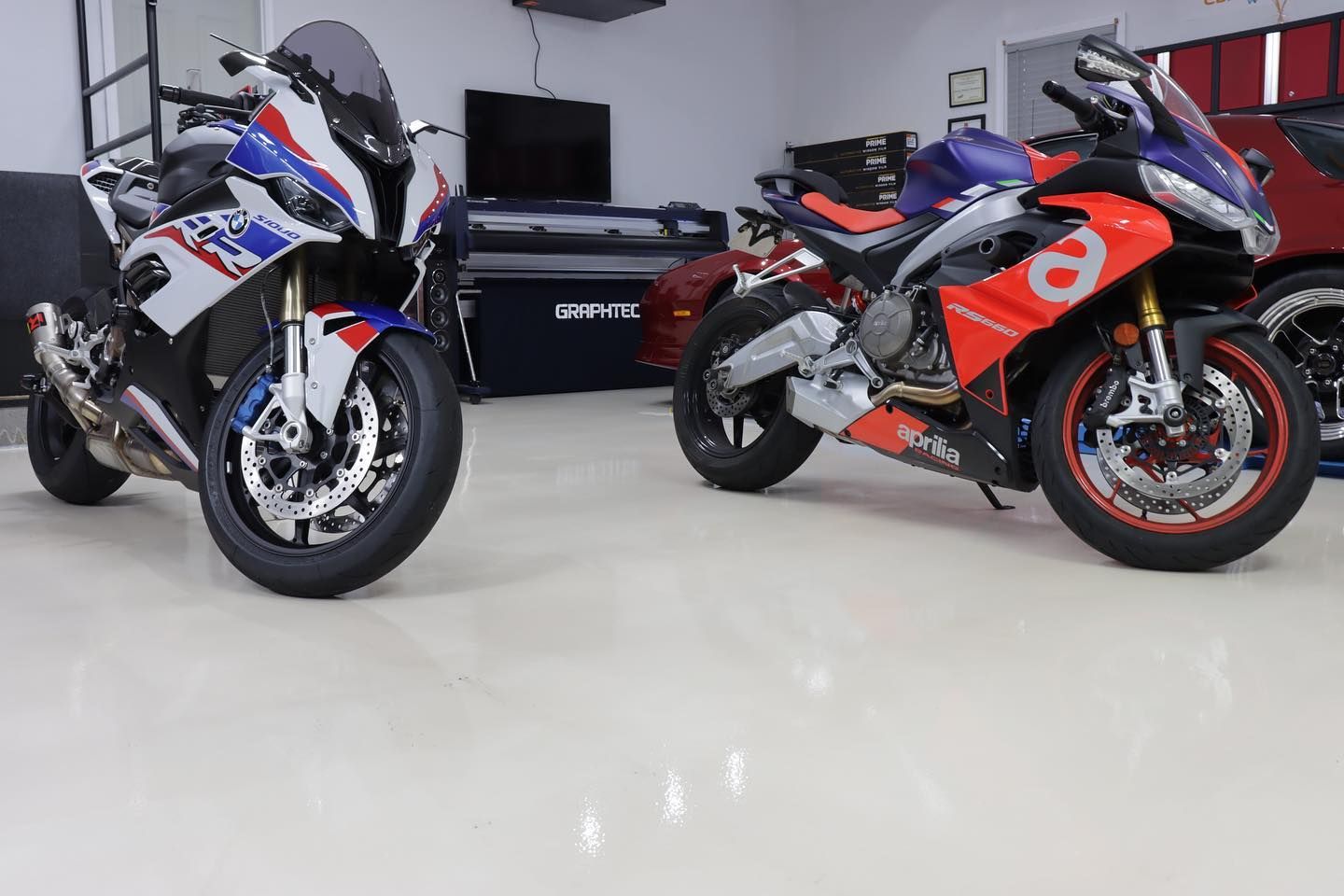 Two sport motorcycles inside a garage, one white and blue, the other red and silver, parked on a shiny floor.