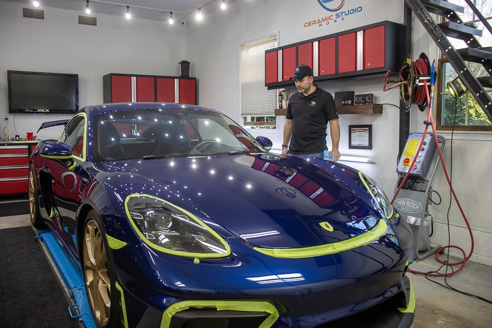 Man inspects a blue sports car in a well-lit garage with red cabinets and tools; the car is partially taped.