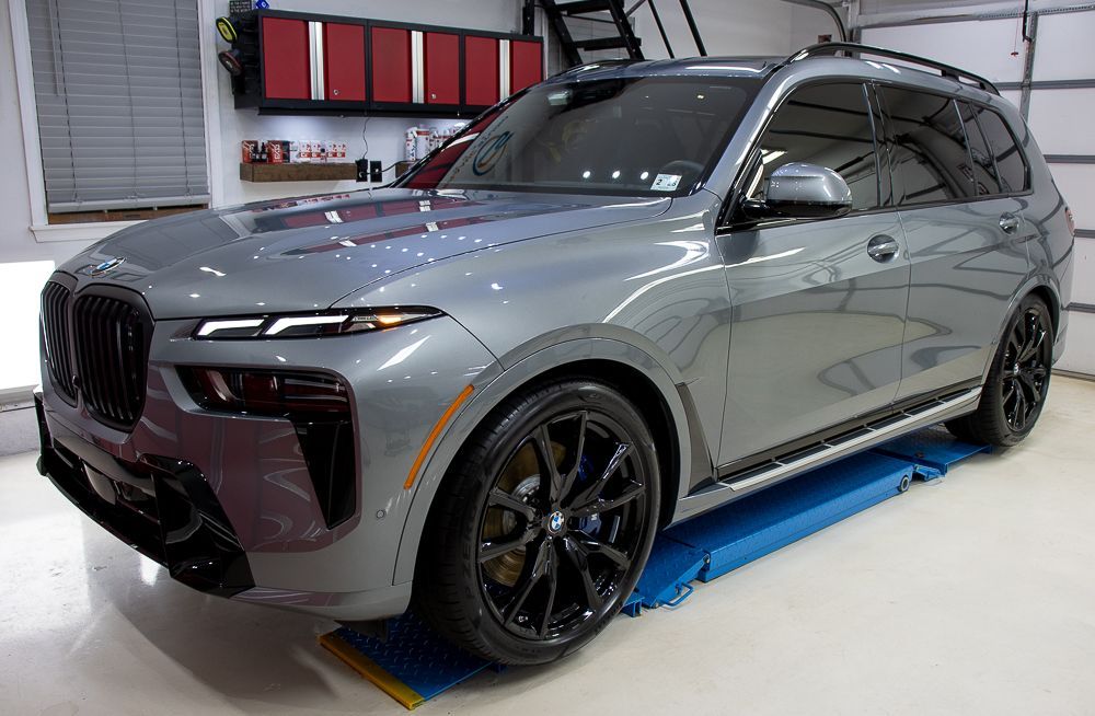 Gray BMW X7 SUV parked inside a garage; black wheels and trim, blue floor mat.