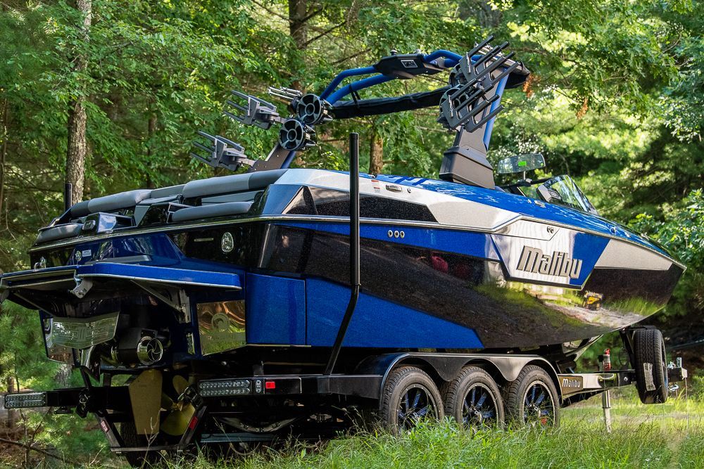Blue and black Malibu wakeboard boat on a trailer, parked in a wooded area.