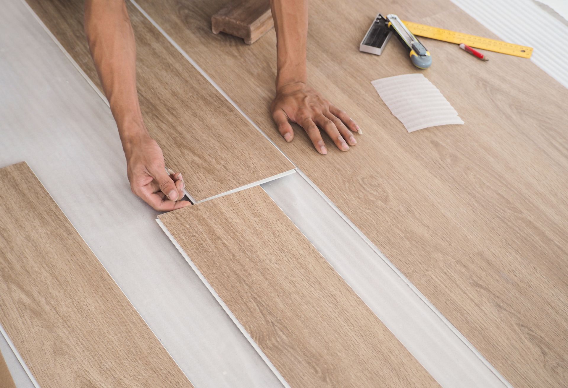 Hands installing a light wood floor plank with tools nearby during home renovation