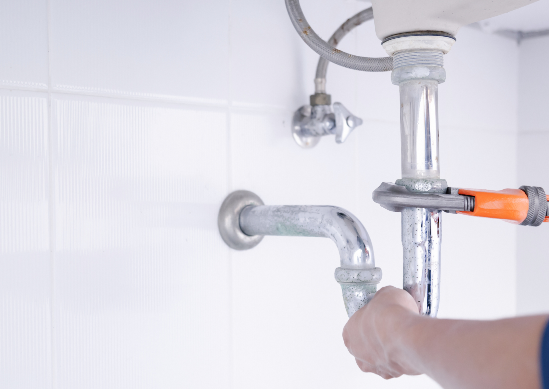 Plumber using a wrench to repair a sink drain pipe in a white bathroom.