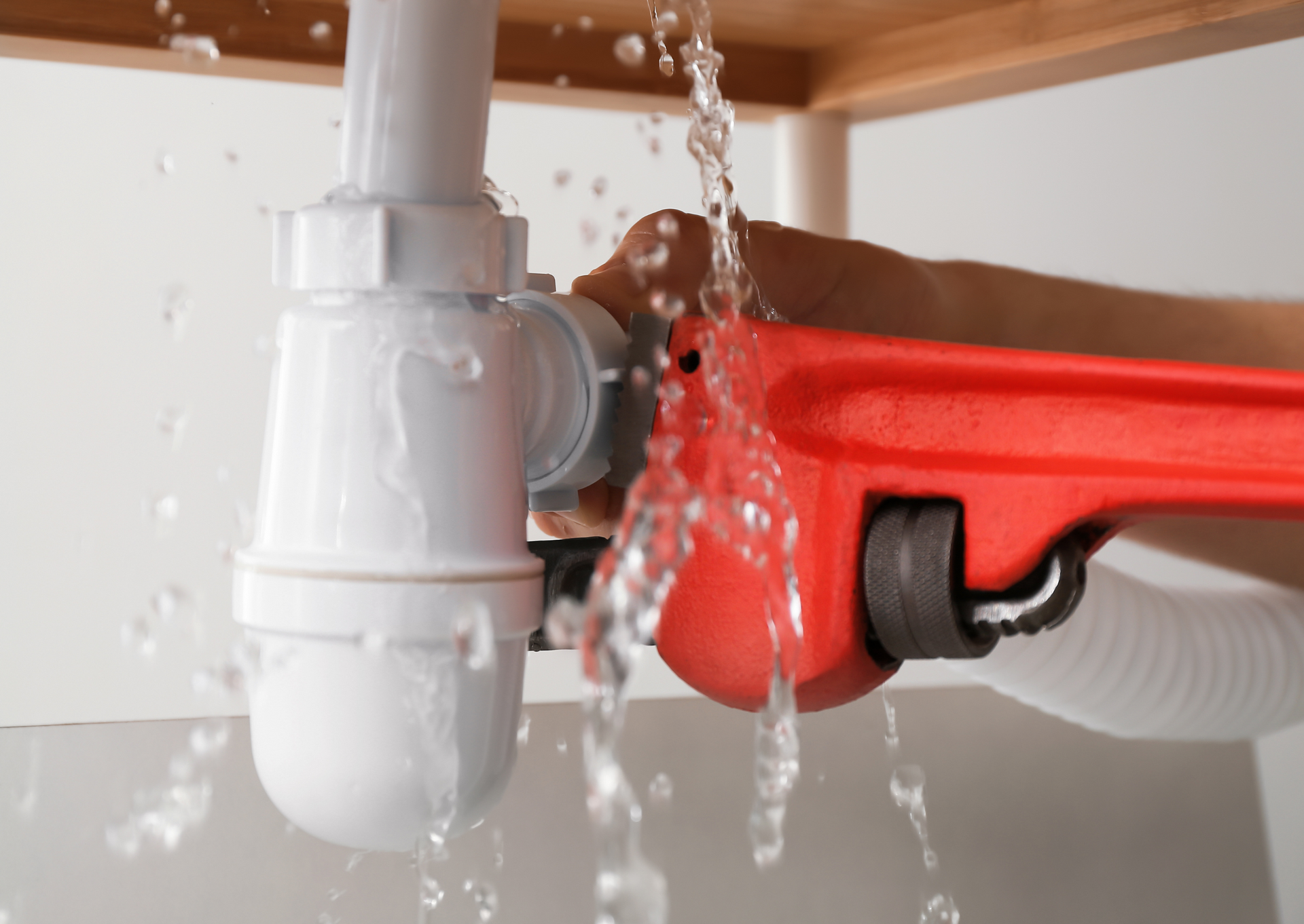 A person tightening a red wrench on a leaking sink pipe, water spraying.