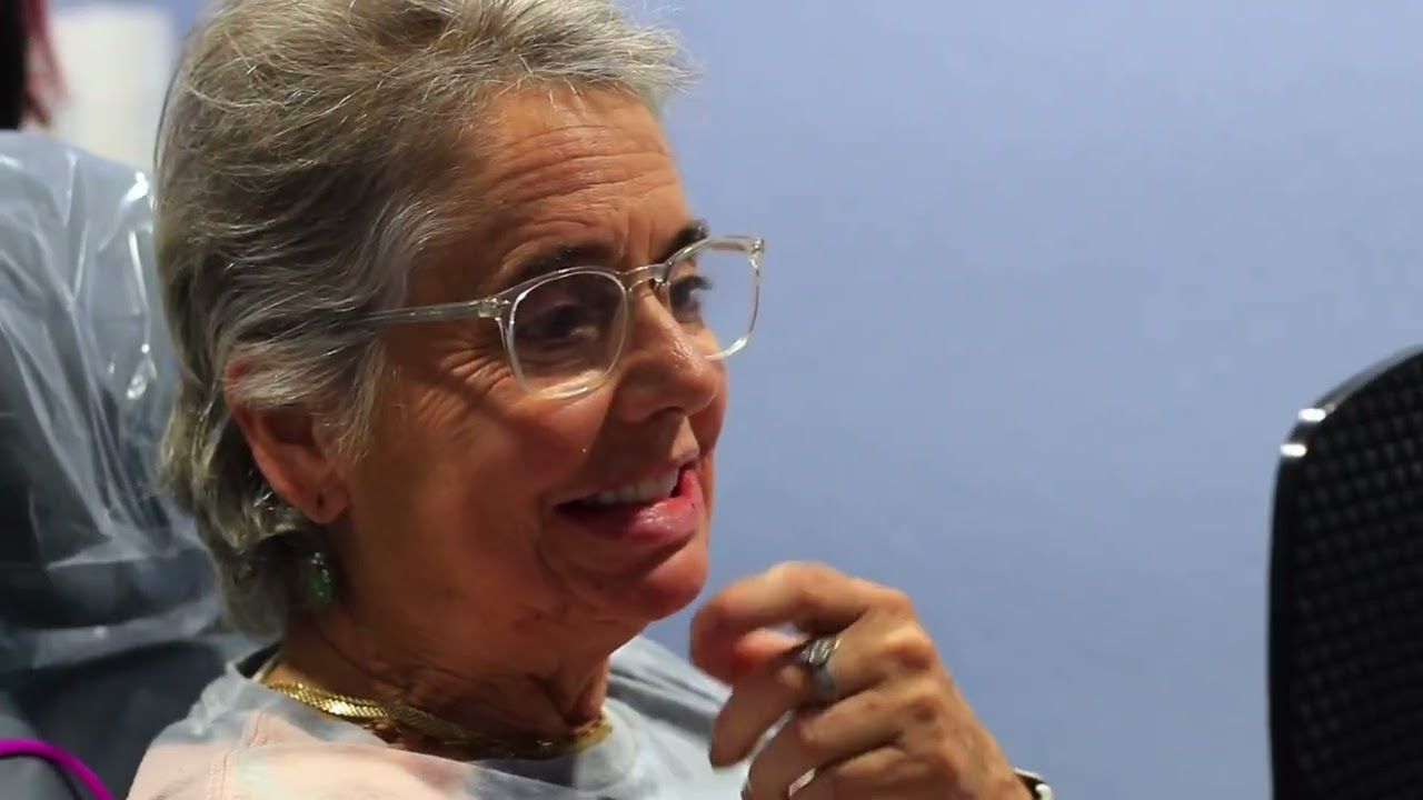 A person with grey hair and clear-framed glasses gestures with one hand while looking toward the right.