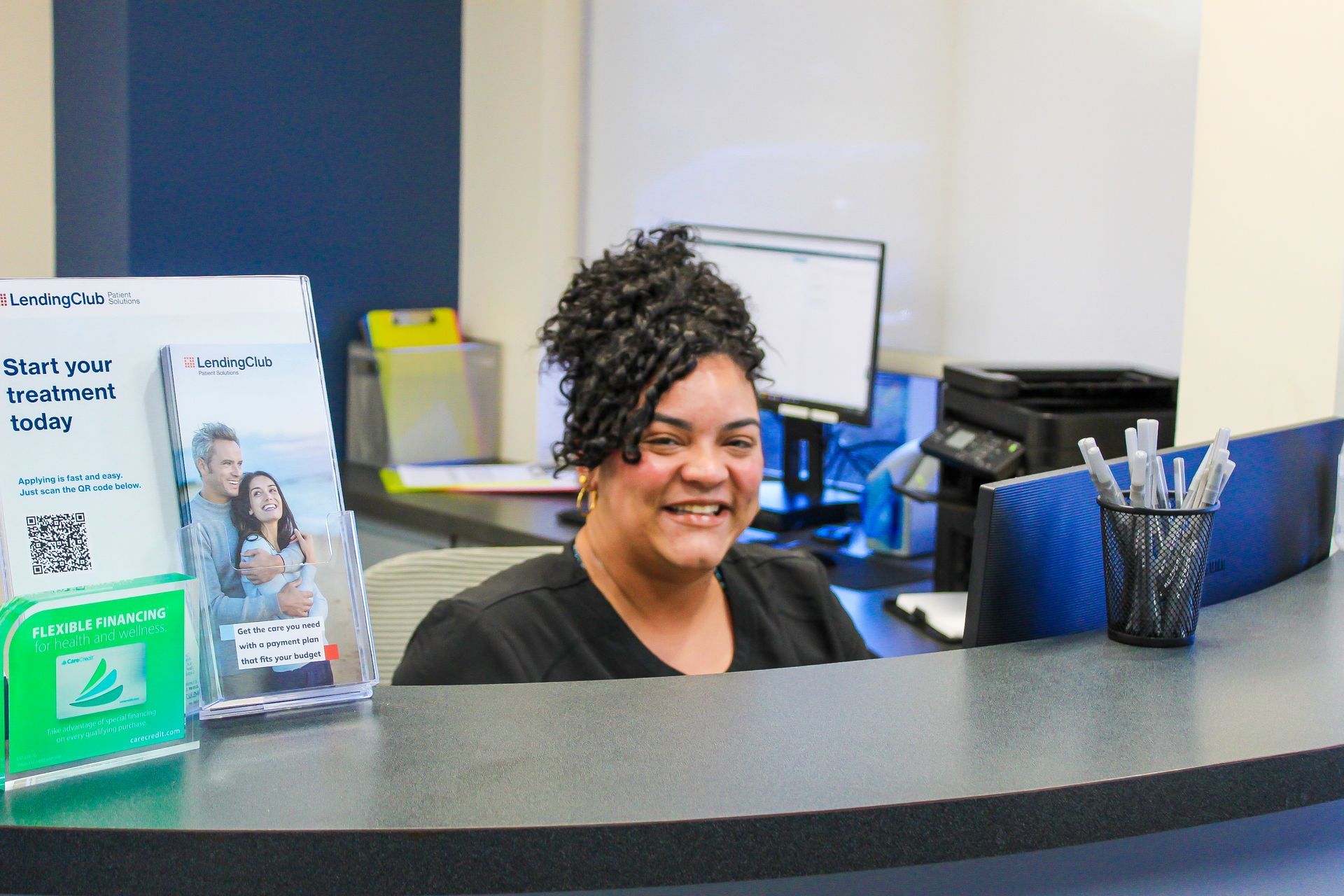 Smiling receptionist at a front desk with brochures and office supplies nearby