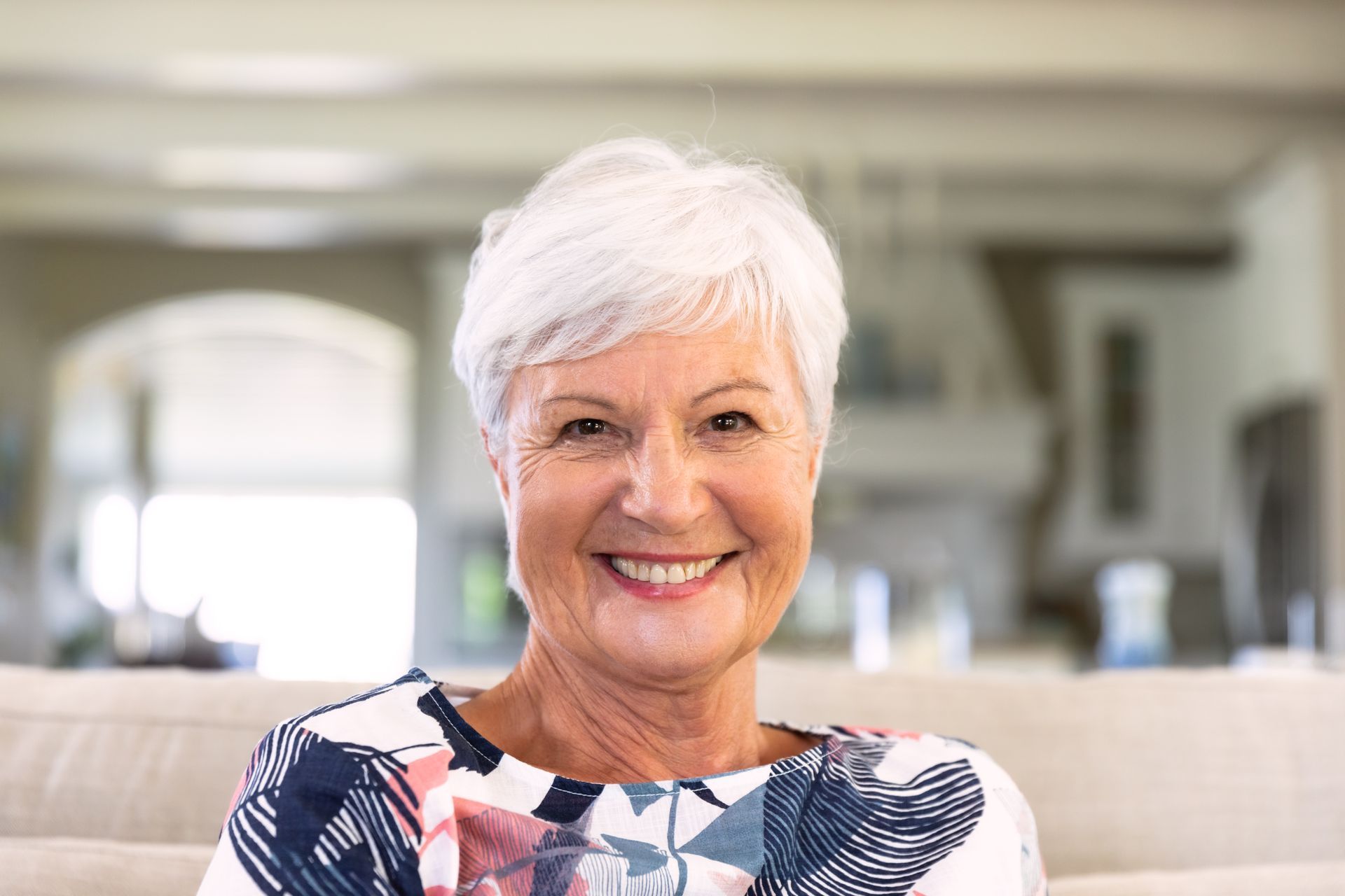 Smiling person with short white hair sitting on a sofa in a bright living room