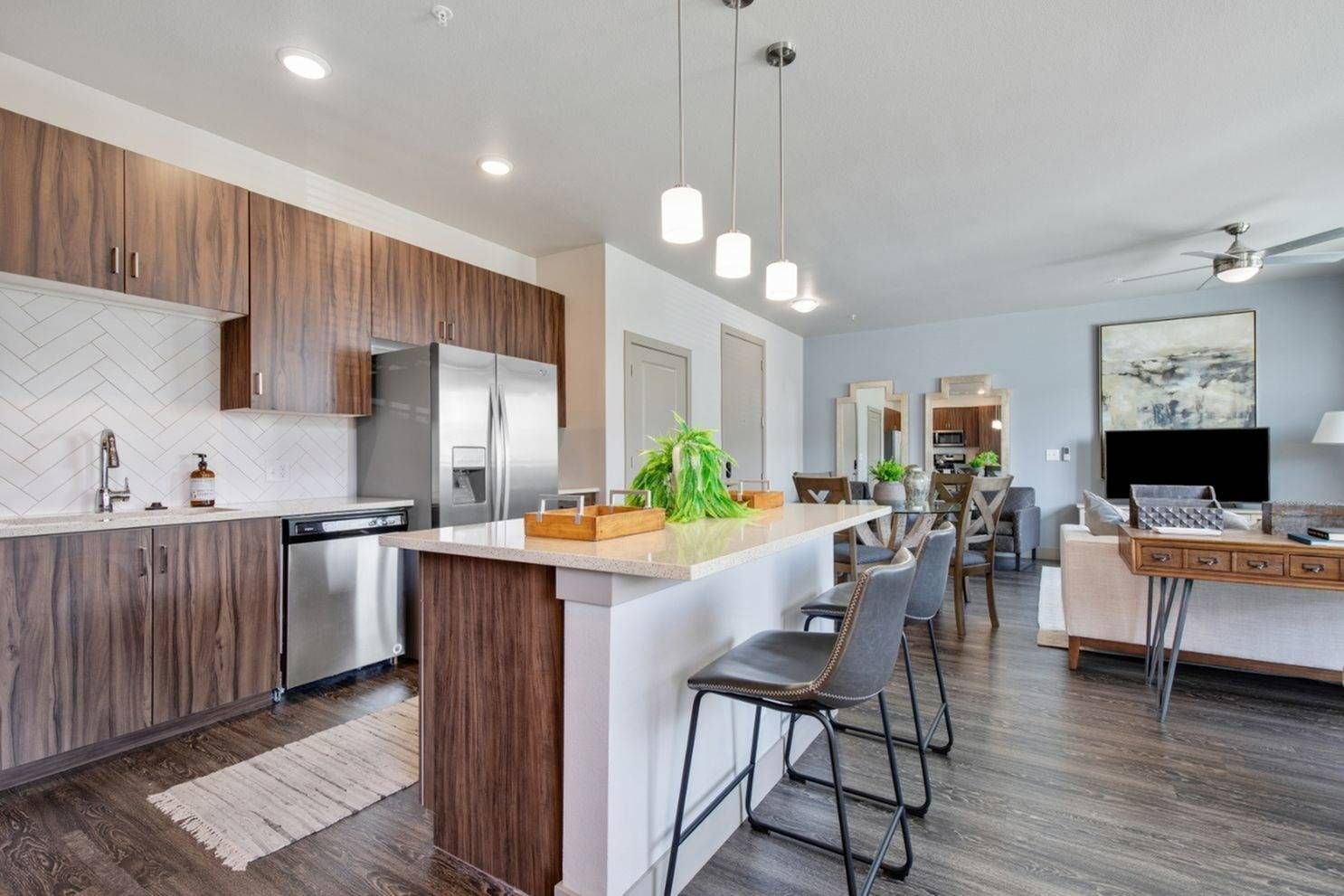 Modern apartment with wood-grain cabinets, stainless steel appliances, island bar, and open living area with fan and artwork.