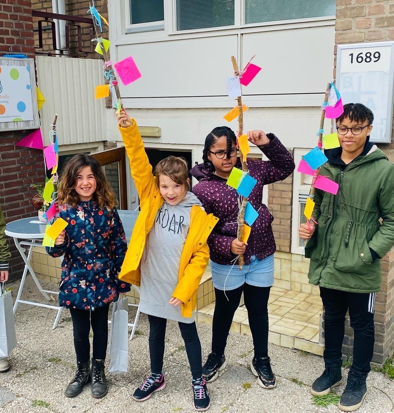 Vier kinderen houden kleurrijke, vlagachtige versieringen vast voor een gebouw met een bordje 