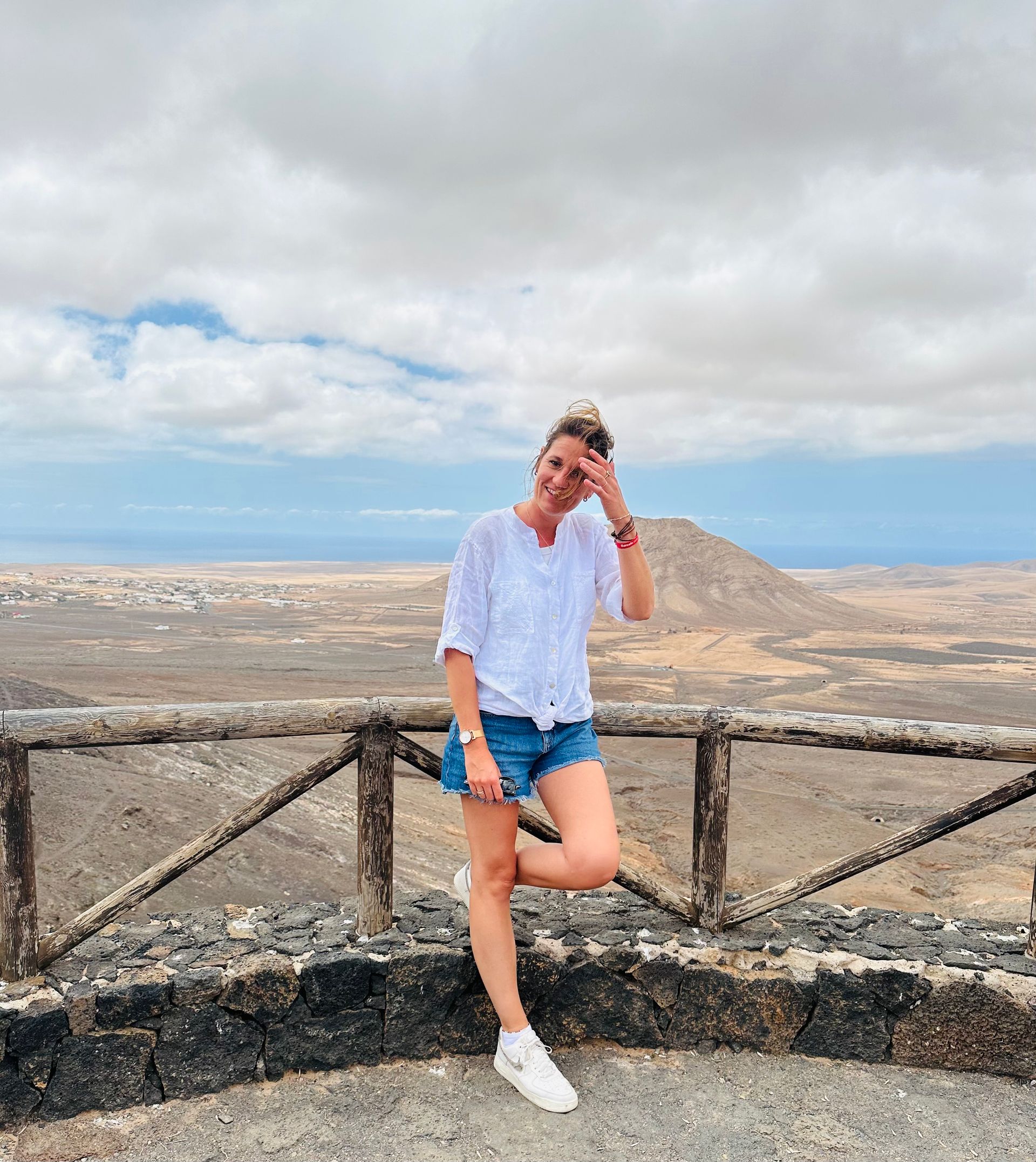 Een vrouw in een witte top en spijkerbroekje leunt glimlachend tegen een reling. Ze kijkt uit over een woestijnlandschap onder een bewolkte hemel.
