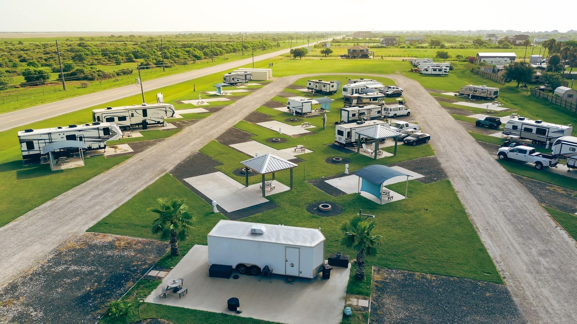 RV park with numerous recreational vehicles parked on grassy lots, surrounded by trees and a roadway.