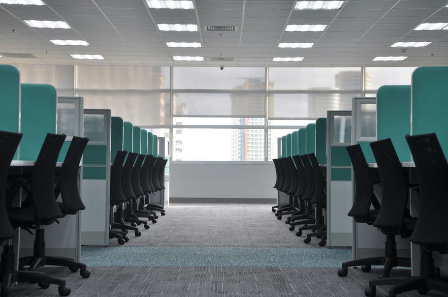An empty office space with two rows of cubicles facing each other, teal partitions, and black rolling chairs.