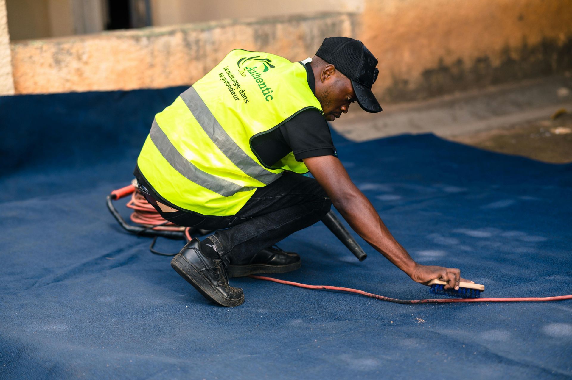 A worker in a high-visibility yellow vest kneeling on a blue surface and scrubbing it with a brush.