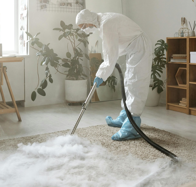 A person in protective gear uses a professional cleaning wand to steam-clean a carpet in a bright, modern living room.