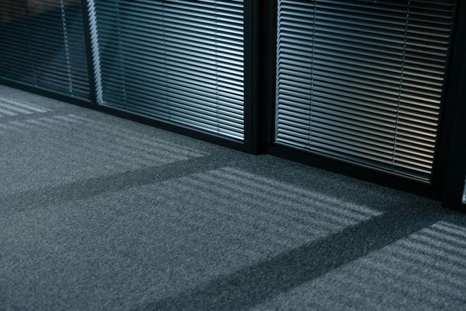 Light filters through closed window blinds, casting striped shadows across a grey carpeted office floor.