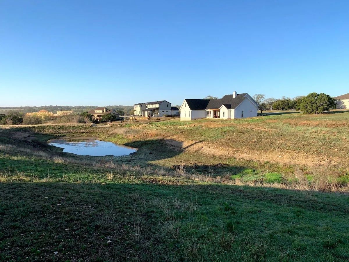 A rural landscape with a pond, houses, and grassy fields under a blue sky.