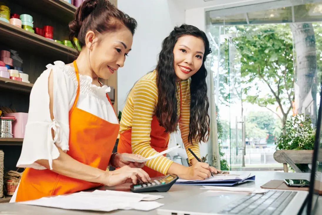 Two women working in a shop, one using a calculator, the other writing.