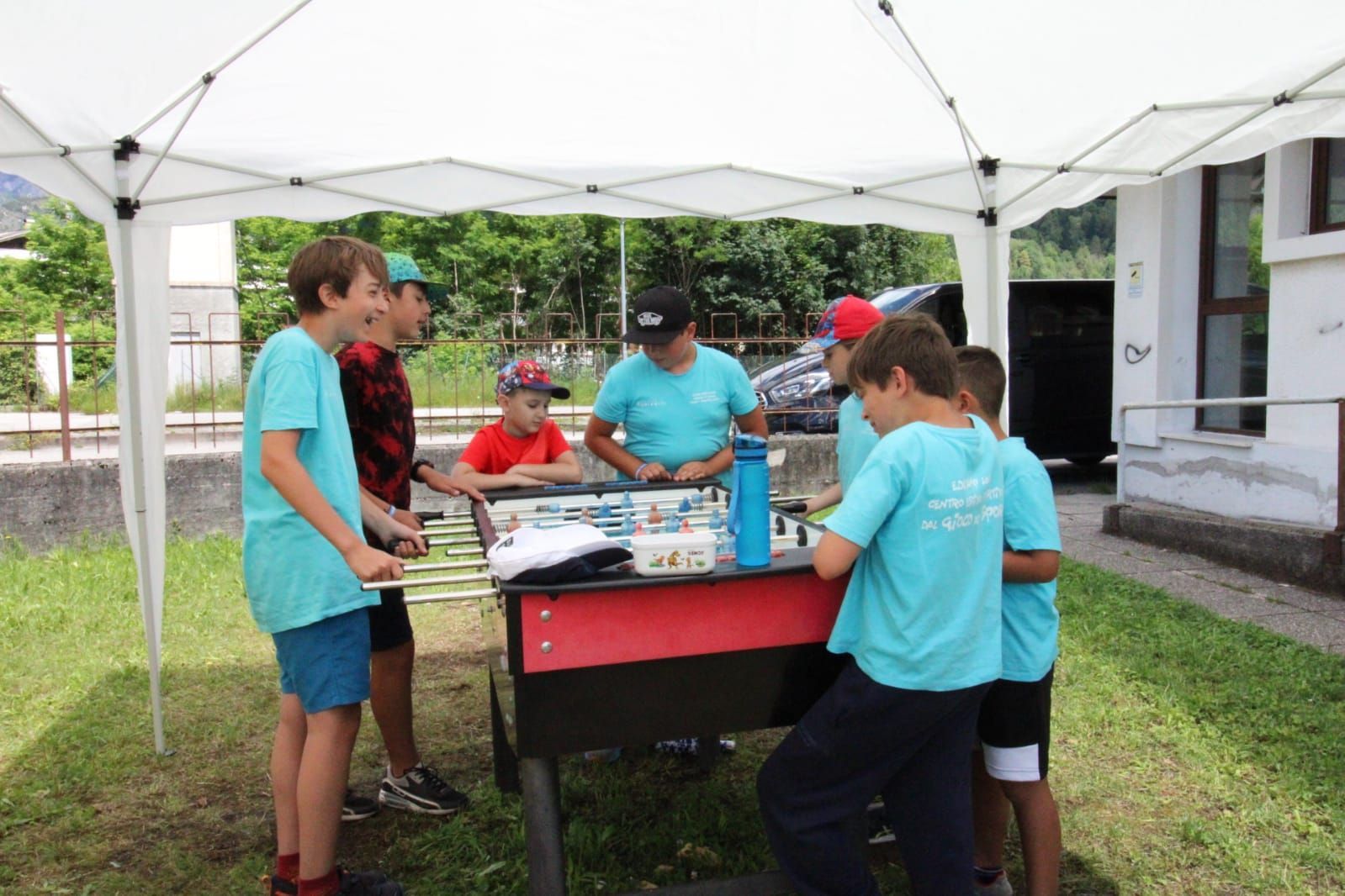 Un gruppo di ragazzi è in piedi attorno a un tavolo da calcio balilla sotto una tenda bianca.