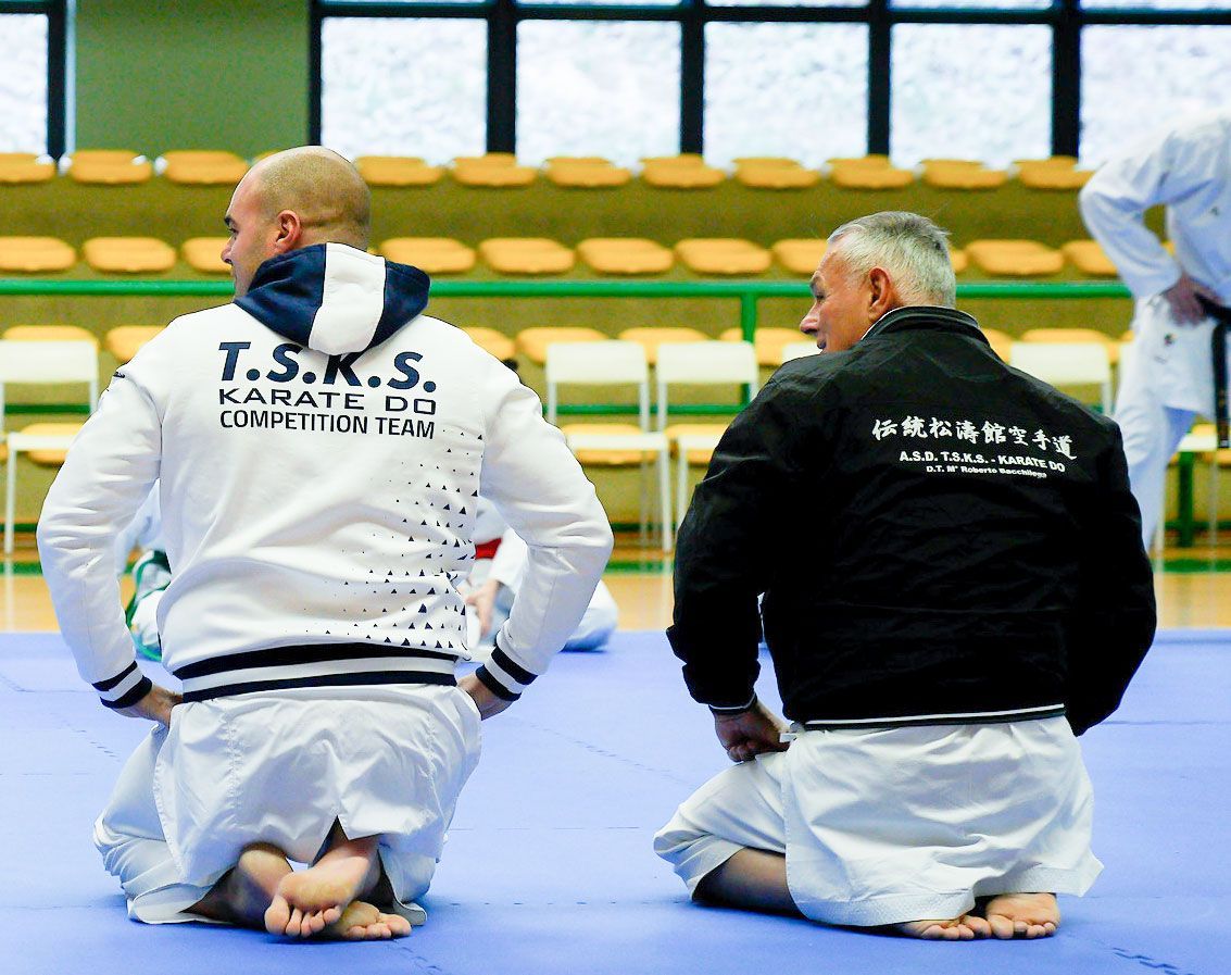 Due uomini sono inginocchiati su una stuoia, uno dei quali indossa una giacca con la scritta