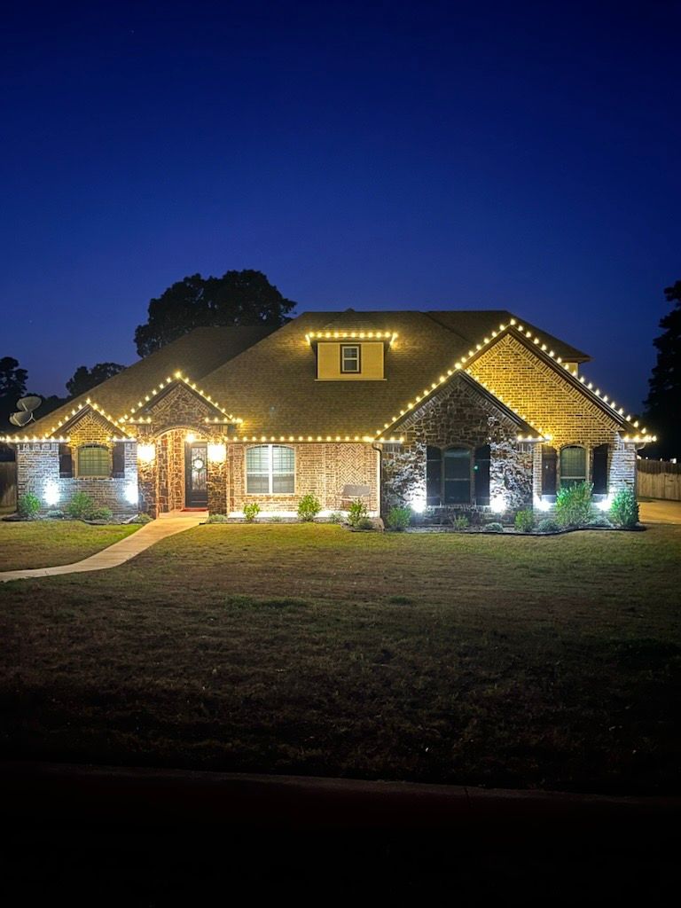 House with warm white lights along the roofline and lit by spotlights at dusk.