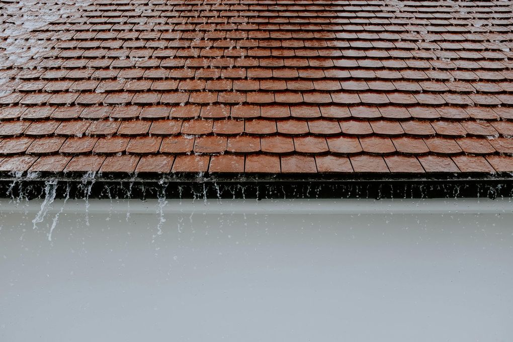 Rain falling off a brown tiled roof into a black gutter, above a white wall.