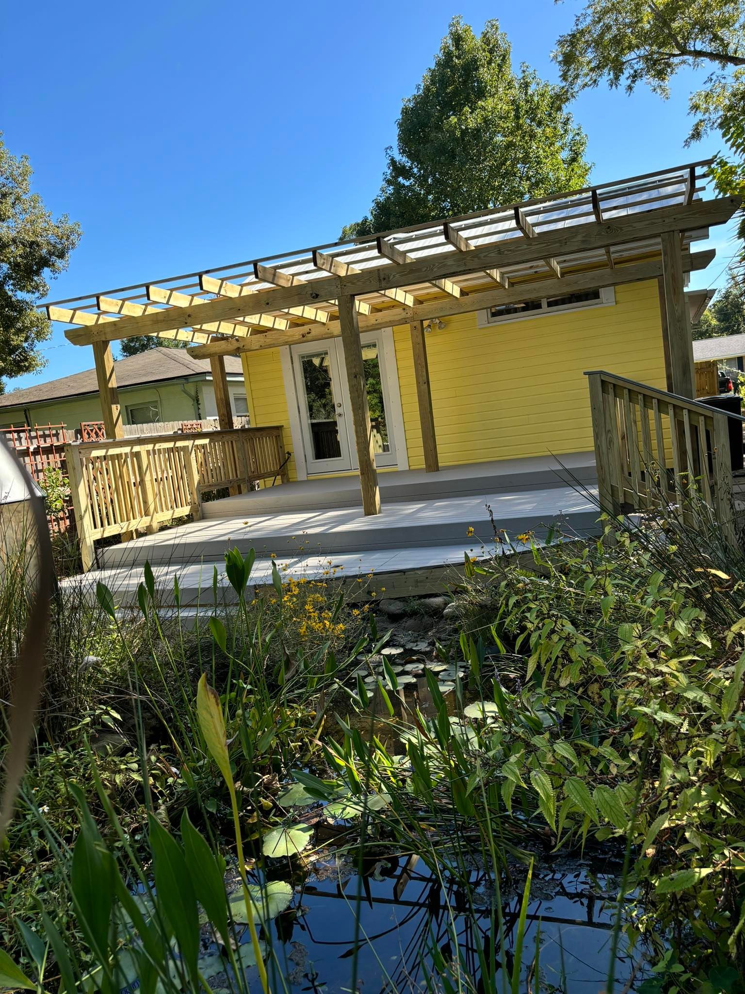 Yellow building with pergola, deck, and railing overlooking a pond with green plants and a clear blue sky.