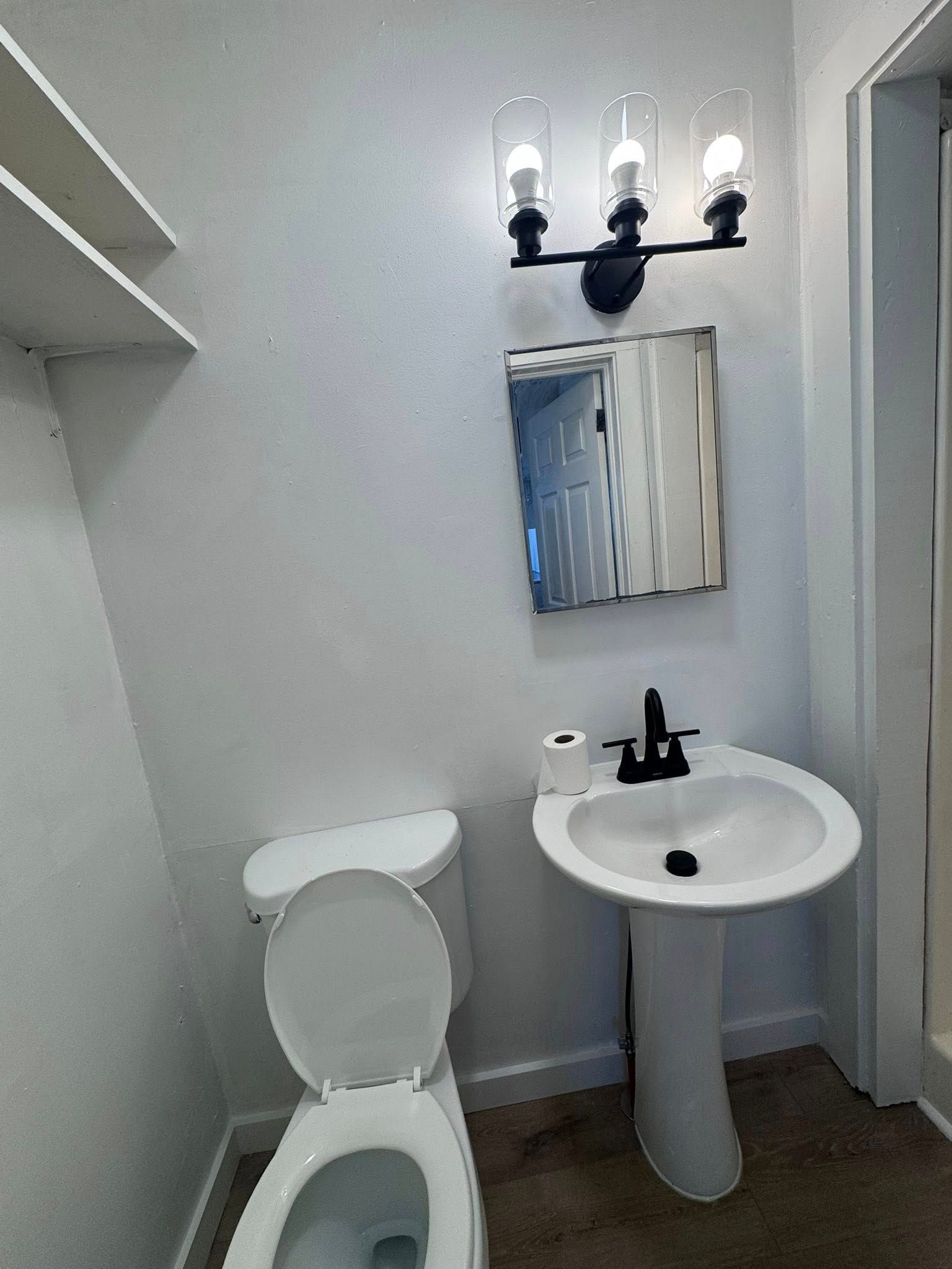 Small bathroom with toilet, sink, and mirror. White walls and a three-bulb light fixture.