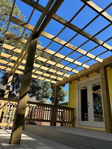 Wooden pergola over a yellow house porch with double doors, bright blue sky in background.