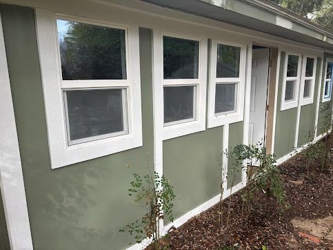 Exterior wall with white-framed windows and a white door; sage green siding. Brown mulch and plants at the base.