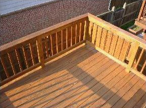 Wooden deck with railing, sunlight, and a view of a brick building.