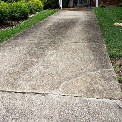 Concrete driveway with dark stains and cracks, flanked by green grass and shrubbery.