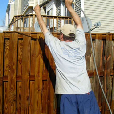 Man power washing a wooden fence outdoors; wearing cap and blue shorts.