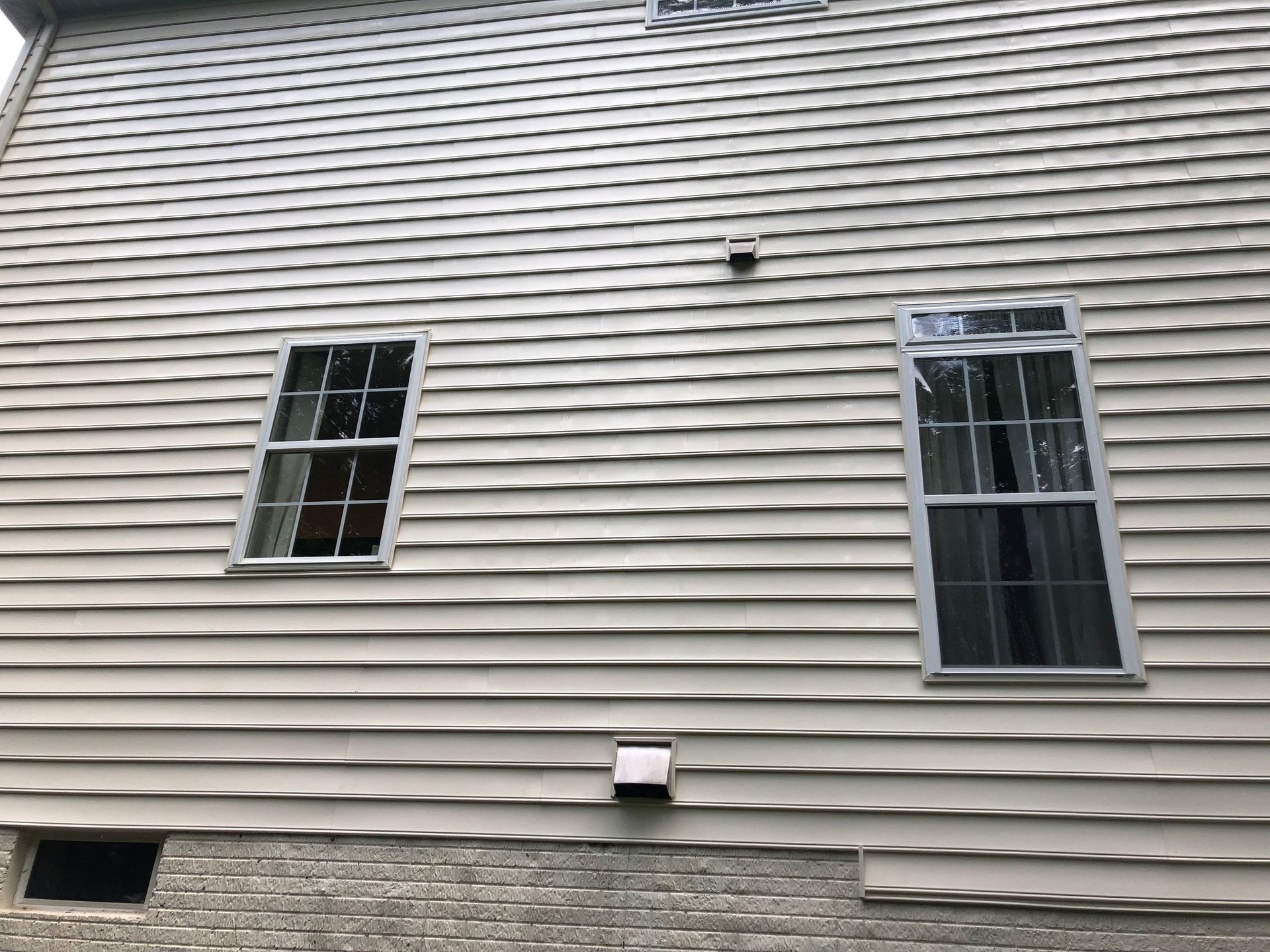 Side of a house with gray wavy siding, two windows, and brick foundation.