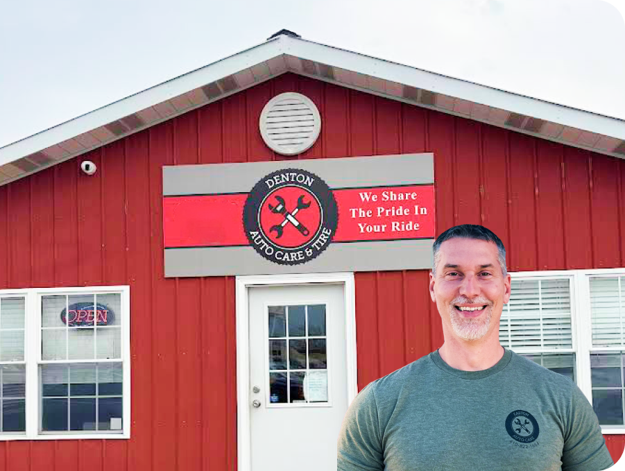 Smiling man stands before a red building with a circular sign and white door. | Denton Auto Care And Tire
