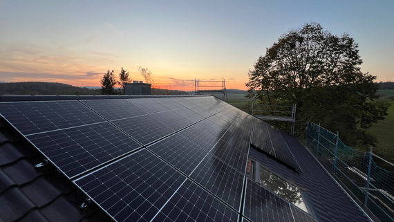 Solar panels on a rooftop at sunset with a silhouetted tree and orange sky.