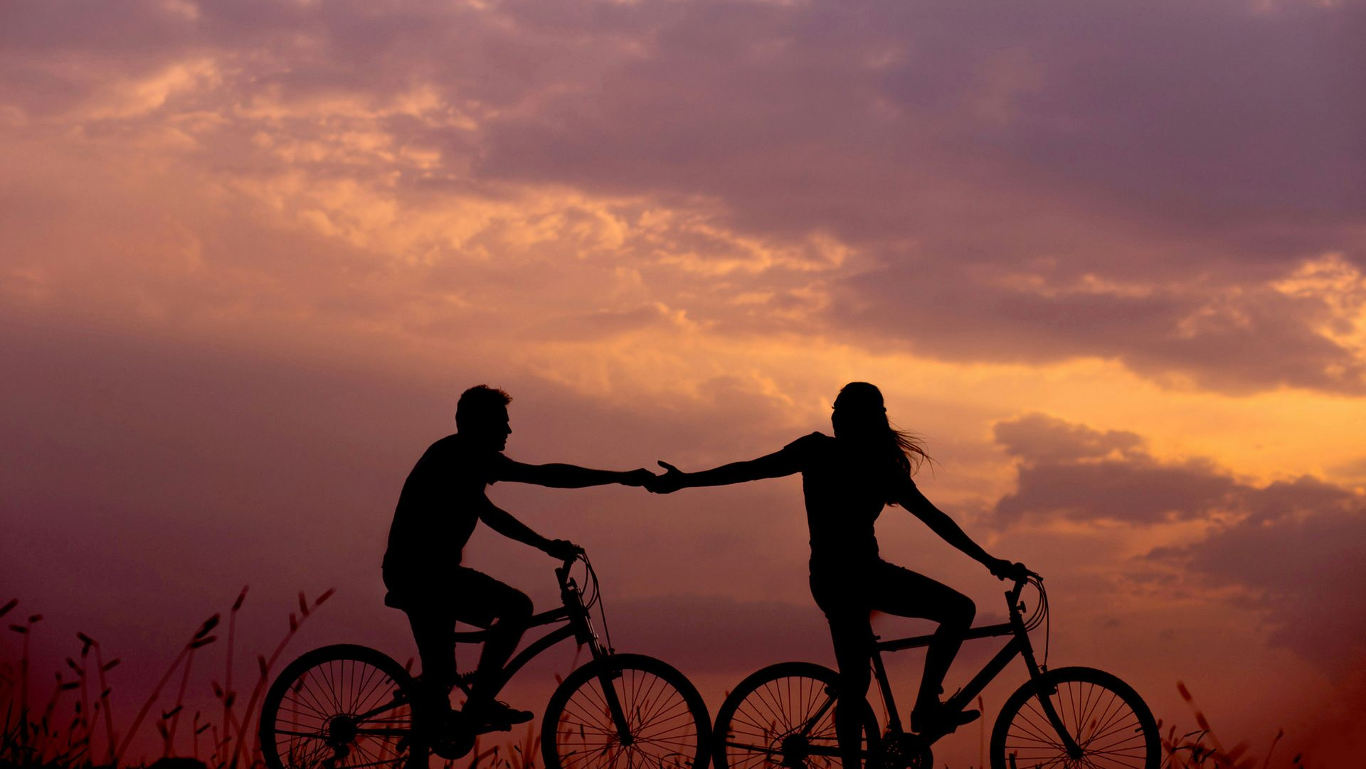 Two cyclists silhouetted at sunset, reaching toward each other on bikes against a colorful sky.